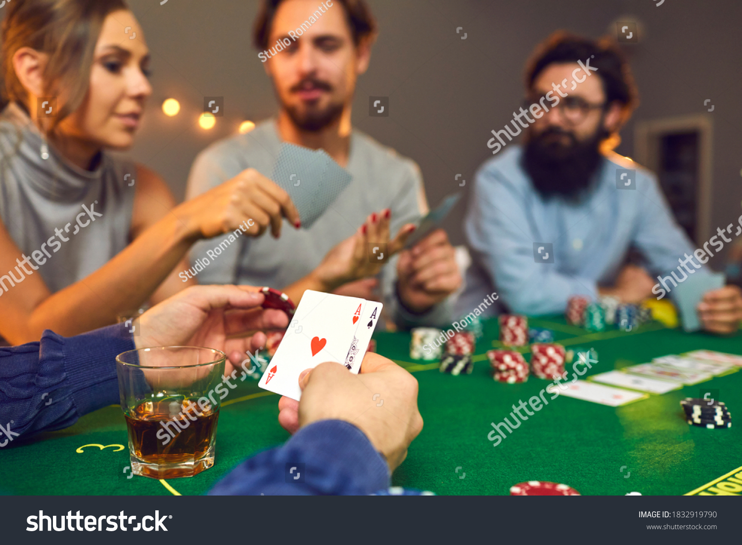 Cropped shot of lucky man holding a chip and pair of aces, hearts and spades, in hand while playing poker sitting at baize card table with friends on casino night, view over shoulder. Gambling concept